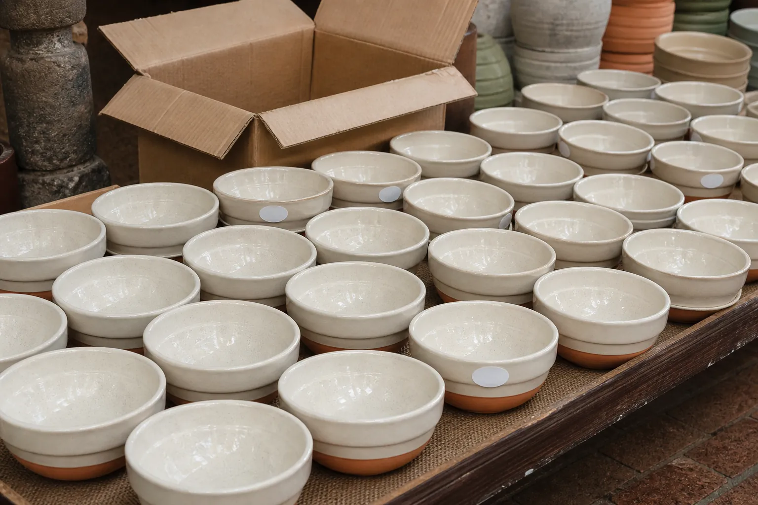 Long visitor-market tray of identical glazed bowls in repeating rows, factory stickers still on a few bases, harsh overhead stall light, a dusty carton half-open behind the display, 1600x900